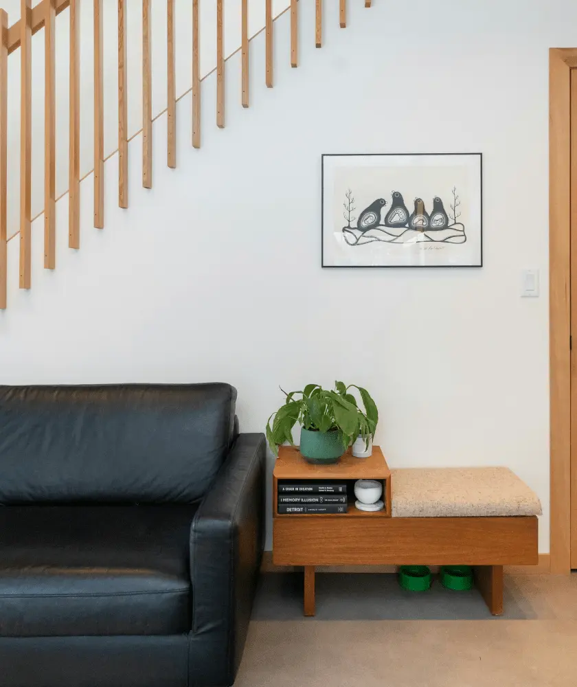 A cozy interior shot shows a black leather sofa next to a light wood open-shelf bench with a plant and books, below a light wood slatted staircase and a framed bird artwork.