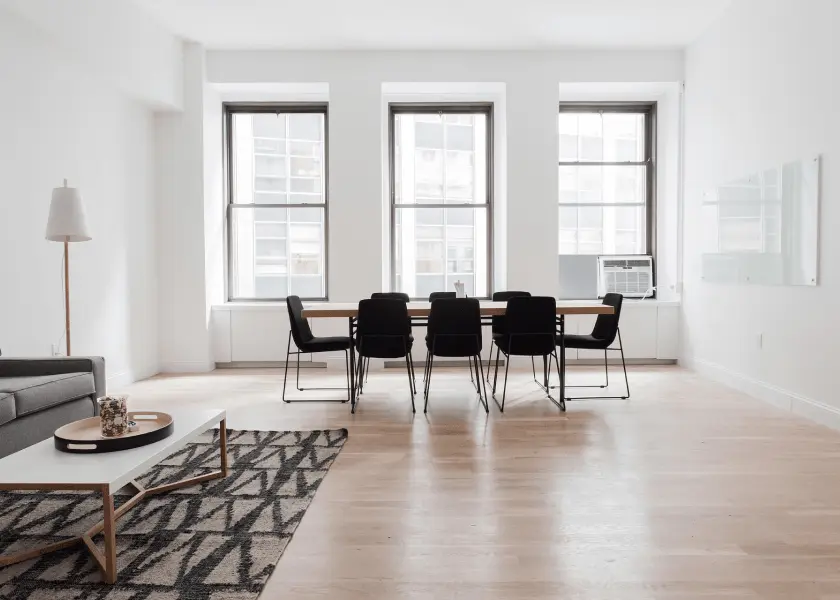 A spacious, minimalist room with light wood floors features a long dining table with black chairs in front of three tall windows, and a living area with a gray sofa and white coffee table on a patterned rug.