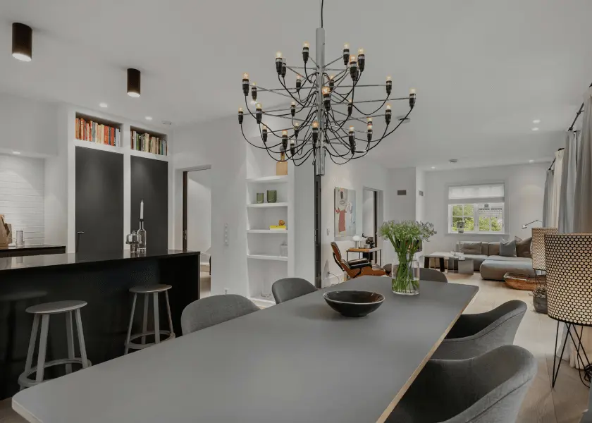A long gray dining table with chairs in a modern home, leading into an open-plan living area. A large, multi-armed chandelier hangs above the table